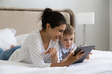 Caring Smiling Young Indian Woman Using Digital Computer Tablet Applications With Happy Small Preschool Kid Son, Playing Games Resting On Bed, Parental Control, Tech Addiction Concept.