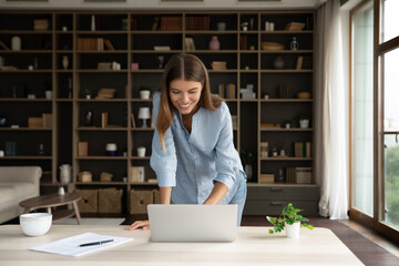 Happy millennial employee girl using laptop, standing at desk with computer, smiling at screen, typing, reading email, chatting online, making video call. Young business woman working frim home office