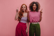 Funny Young Interracial Girls Point Index Fingers Up With Copy Space. European Redhead Lady In Tight Top And Trousers Poses With African Brunette With Voluminous Hairstyle In T-shirt, Loose Pants.