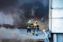 firefighters are standing on roof. Firefighters view from afar. Two employees of fire department. Concept - fighting fire in industrial buildings. Firefighters on background of black smoke.