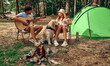 A young family with a child and a labrador dog sit by the campfire near the tent, play the guitar in the pine forest. Camping, recreation, hiking.