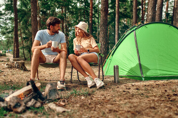 A loving young couple went on a weekend for a picnic, sit near a campfire and a tent, drink coffee in a pine forest. Camping, recreation, hiking.