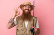 Young caucasian ginger fisherman with long beard holding a rod isolated on pink background cheerful and confident showing ok gesture.