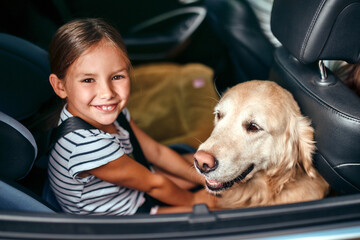 Wall Mural - Cute little girl child in a car seat protected by seat belts together with her friend dog labrador going on weekend. Leisure, travel, tourism.