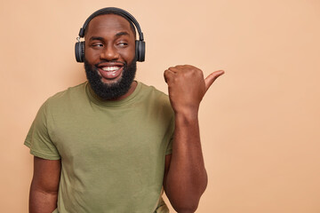 Wall Mural - Horizontal shot of optimistic dark skinned adult man points away on blank space shows place for your advertisement listens audio track in headphones has happy mood isolated over beige background