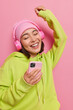 © wayhome.studio  - Photo of cheerful teenage girl with Asian appearance dances with rhythm of music listens favorite song in headphones from playlist holds mobile phone moves actively wears sweatshirt hat poses indoor