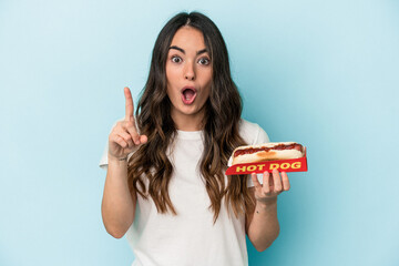 Young caucasian woman holding a hot dog isolated on blue background having an idea, inspiration concept.