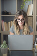 © simona - Attractive smiling young woman wearing eyeglasses using laptop while sitting in front of table at home, Beautiful woman using laptop. Smiling girl browsing social media content or attending online