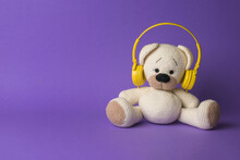 A White Knitted Bear With Yellow Headphones On A Purple Background.