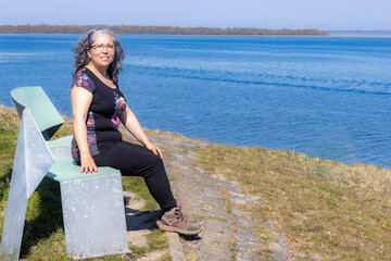 Smiling mature Latin woman sitting on a bench on a hill with Lake Grevelingen in the background, black clothes, grayish curly hair, glasses, tanned skin, sunny day in Zonnemaire, Zeeland, Netherlands