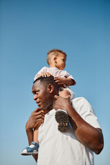 Wall Mural - Vertical portrait of happy African-American father carrying son on shoulders against blue sky while enjoying walk in sunlight