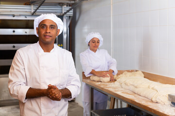 Wall Mural - Portrait of confident hispanic bakery owner in white uniform posing against busy workers background