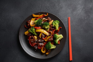 Asian teriyaki beef with red and yellow bell peppers, broccoli and sesame seeds on a plate on the table. Spicy teriyaki beef stir fry with vegetables and rice  on a dark background