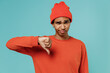 Young sad unhappy displeased african american man 20s in orange shirt hat showing thumb down dislike gesture isolated on plain pastel light blue background studio portrait. People lifestyle concept.