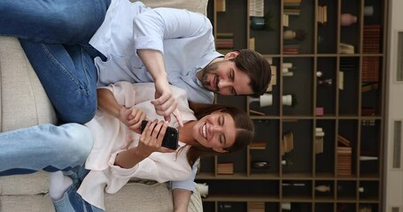 Wall Mural - Young man woman in love hug on couch use phone entertainment app browse funny news at social media talk have fun at home. Family couple enjoy networking with friends online using cell. Vertical video