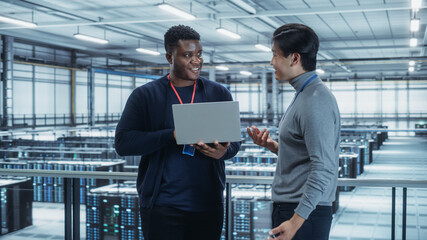 Two Multiethnic Male Data Center System Administrators and IT Specialists Talk, Use Laptop. Information Technology Engineers work on Cyber Security Network Protection in Cloud Computing Server Farm.