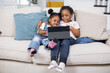 © sofiko14 - Two joyful african girls sitting together on comfy couch and using digital tablet. Pretty little sisters watching cartoons during free time at home.