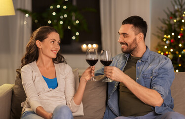 Canvas Print - leisure, clebration and winter holidays concept - happy couple drinking red wine at home in evening over christmas tree lights on background