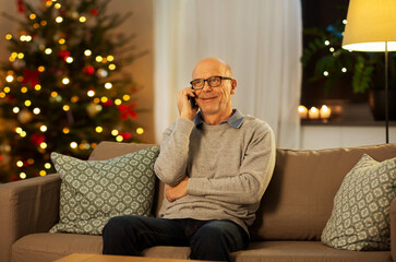 Wall Mural - technology, winter holidays and communication concept - happy smiling senior man calling on smartphone at home in evening over christmas tree lights on background
