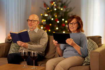 Wall Mural - leisure and winter holidays concept - happy senior couple reading book and using tablet pc computer at home in evening over christmas tree lights on background