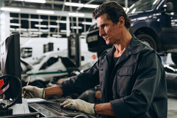 Waist up portrait view of the auto mechanic standing near his computer and checking orders with opened car at the background. Car repair concept