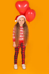 Wall Mural - a little beautiful happy blonde girl in a New Year's jumper and plaid trousers with a Santa Claus hat holding red balloons on a yellow background