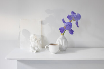 Wall Mural - Blue iris in a corrugated vase with a piece of craft paper on a white table. Figurine of the angel. Cup of tea. Place for text