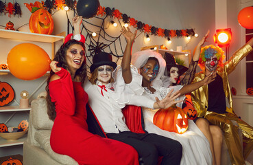 Wall Mural - Happy friends disguised as crazy evil monsters having fun at Halloween party. Adult people with scary makeup sitting together on sofa in living room, looking at camera and waving hello for group photo