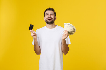 Young handsome happy smiling man holding banking card and cash in his hands isolated on yellow background.