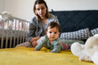 © Miljan Živković - Front view on happy small caucasian baby lying on the bed crawling by her mother at home in day copy space childhood growing up family concept