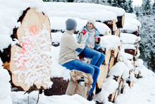 Couple Enjoying Coffee While Sitting On Snow Covered Logs In Forest
