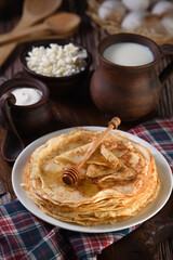 Homemade thin pancakes with honey stacked in a stack, on a wooden table with a mug of milk, a pot of sour cream and eggs in a basket. Traditional Slavonian, pagan holiday (Maslinitsa). Country style f