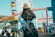 © PR Image Factory - rear view of beautiful asian korean woman wearing straw hat and carrying backpack walking in front of colorful ferris wheel at amusement park on sunny day. girl looking at roller coaster outdoors.