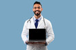 © Prostock-studio - Arabic Male Doctor Showing Laptop Empty Screen Over Blue Background