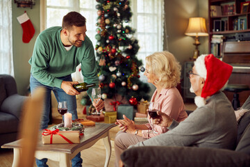 Wall Mural - Happy man serves wine to senior parents while celebrating Christmas together at home.