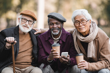 Wall Mural - Smiling elderly interracial friends with cellphone and coffee to go looking at camera outdoors