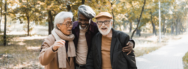 Wall Mural - African american man hugging interracial senior friends in autumn park, banner