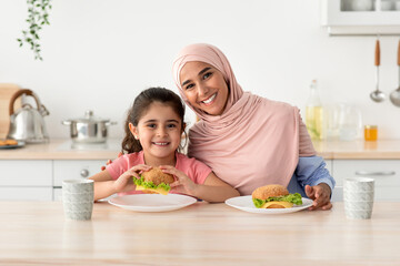 Wall Mural - Happy Muslim Mom In Hijab And Little Daughter Eating Sandwiches In Kitchen