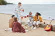 © Seventyfour - Full length portrait of diverse group of friends clinking beer cups on beach in Autumn and laughing