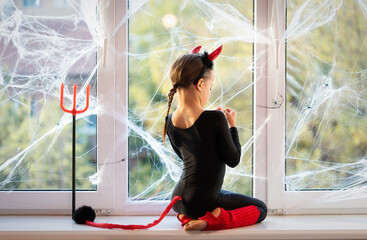 Back view of little girl in costume of little devil sitting on window sill
