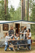 © Seventyfour - Vertical full length view at diverse group of young people clinking beer bottles while enjoying picnic outdoors at campsite with trailer van