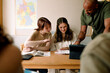 © Maskot - Male professor teaching teenage girls at desk in high school classroom