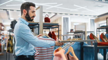 Wall Mural - Handsome Male Clothing Store Assistant Works in Fashionable Shopping Mall. Professional Shop Sales Retail Assistant Hangs New Colorful Collection with Sustainable Casual Design on Display Racks.