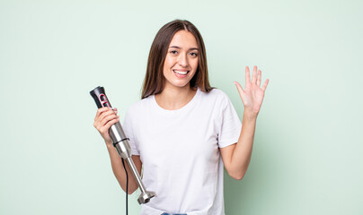 Poster - young pretty woman smiling happily, waving hand, welcoming and greeting you. hand mixer concept