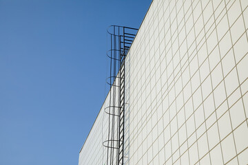 Long metal staircase on the gray modern facade of an industrial building, warehouse or shopping center against the blue sky. Symbol of the stairway to heaven