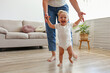 First steps of a little girl. A blond toddler learning to walk at home with the help of loving mother. Close up, copy space for text, background.