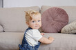 © Evrymmnt - One year old child learning to stand up at living room. Interior background. Adorable blonde little girl in denim overalls leaning on the couch at home. Close up, copy space for text.