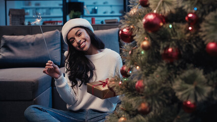 Wall Mural - cheerful african american woman in santa hat holding sparkler and present near blurred christmas tree