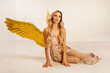 A beautiful young model with big golden angel wings is sitting in the studio. white background