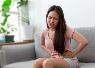 Wall Mural - Asian young woman was holding her hand to catch her belly, she has a stomach pain on the sofa at home.